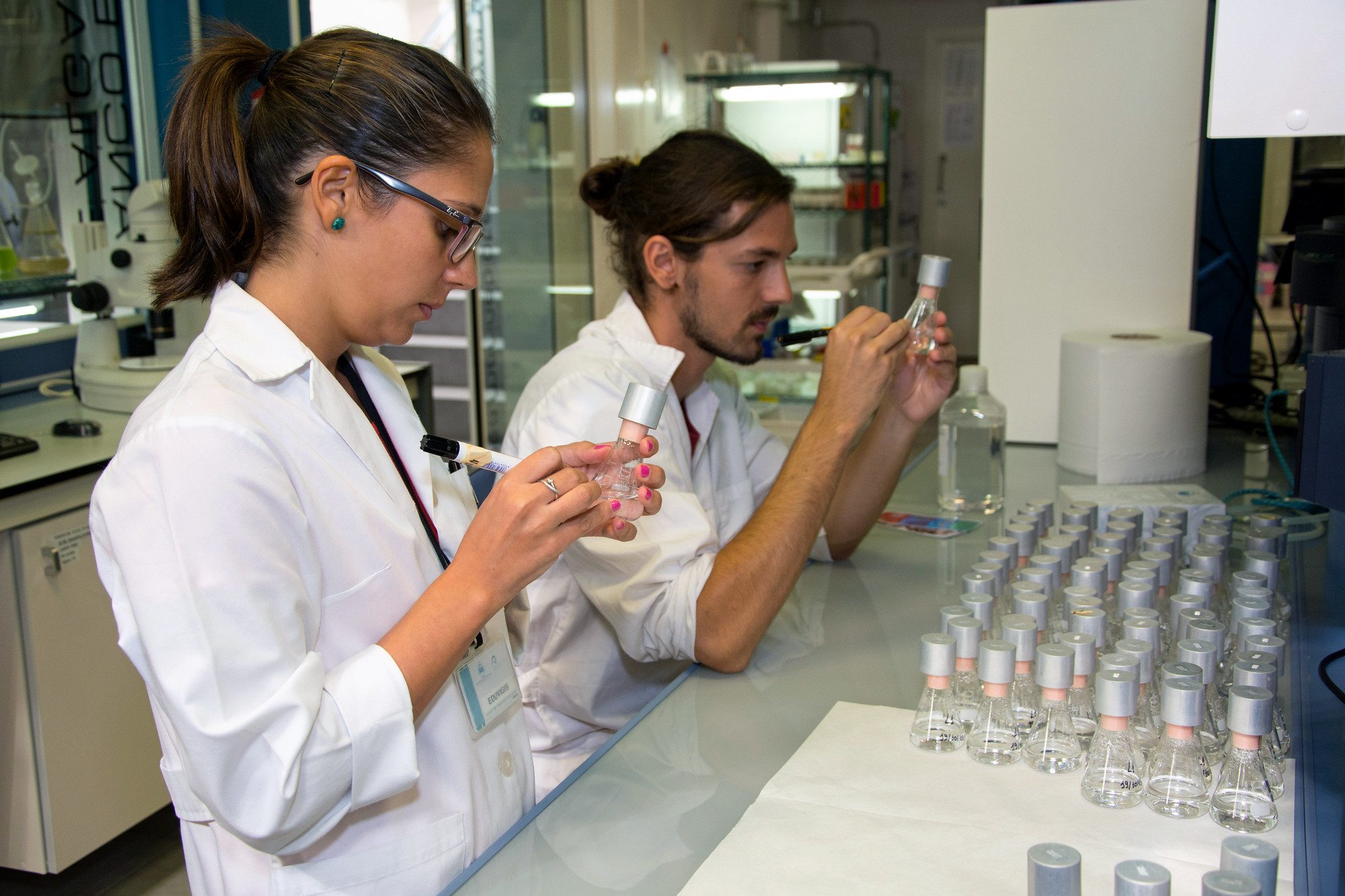Una mujer y un hombre jóvenes, con bata blanca, en un laboratorio, ante una mesa con numerosos recipientes de muestras, escriben con rotulador en una de ellas.